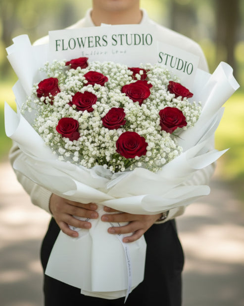 A bouquet of red roses and white baby's breath flowers wrapped in white paper, with printed cards reading 'FLOWERS STUDIO' and 'STUDIO' visible behind the arrangement.