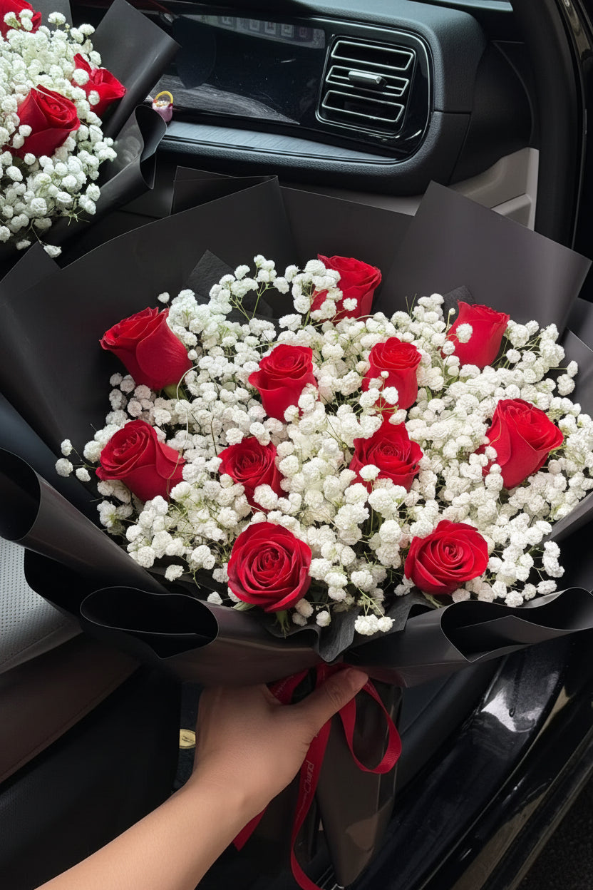 A large bouquet of red roses and white baby's breath wrapped in black paper, tied with a red ribbon, placed inside a car. A small card is attached to the bouquet.