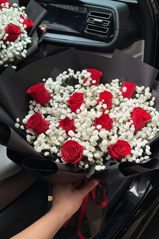 A large bouquet of red roses and white baby's breath wrapped in black paper, tied with a red ribbon, placed inside a car. A small card is attached to the bouquet.