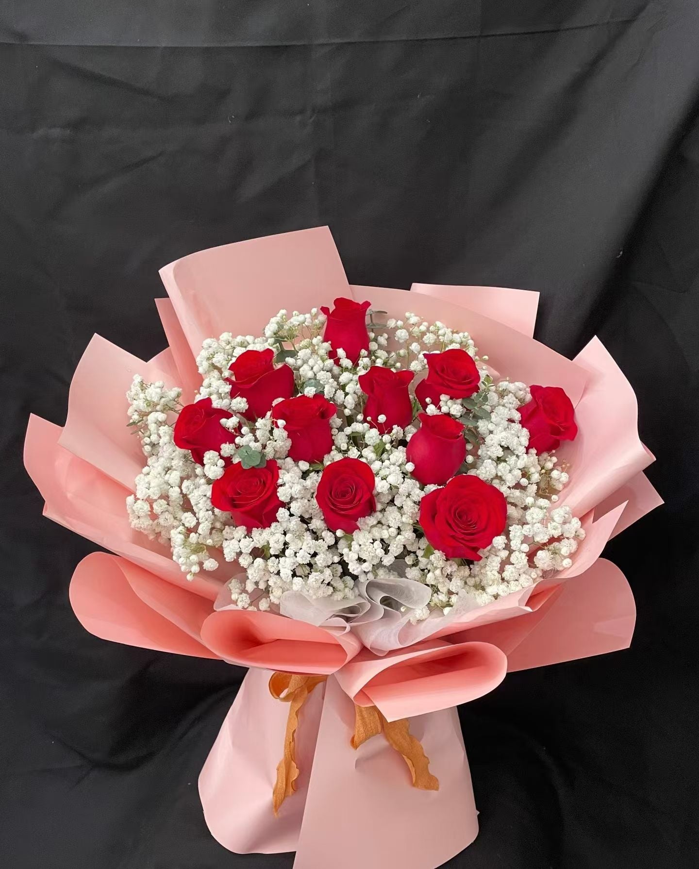 A bouquet of 11 red roses mixed with white baby's breath, wrapped in pink paper with a pink ribbon bow, set against a black fabric background.