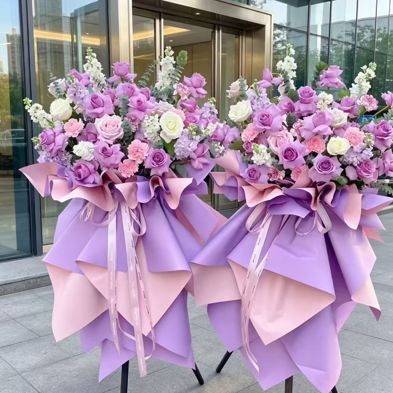 Two large floral welcome or congratulations baskets decorated with pink, purple, and white roses, hydrangeas, and green foliage, wrapped in matching pink and purple paper with ribbon bows, standing outdoors in front of a glass building entrance.