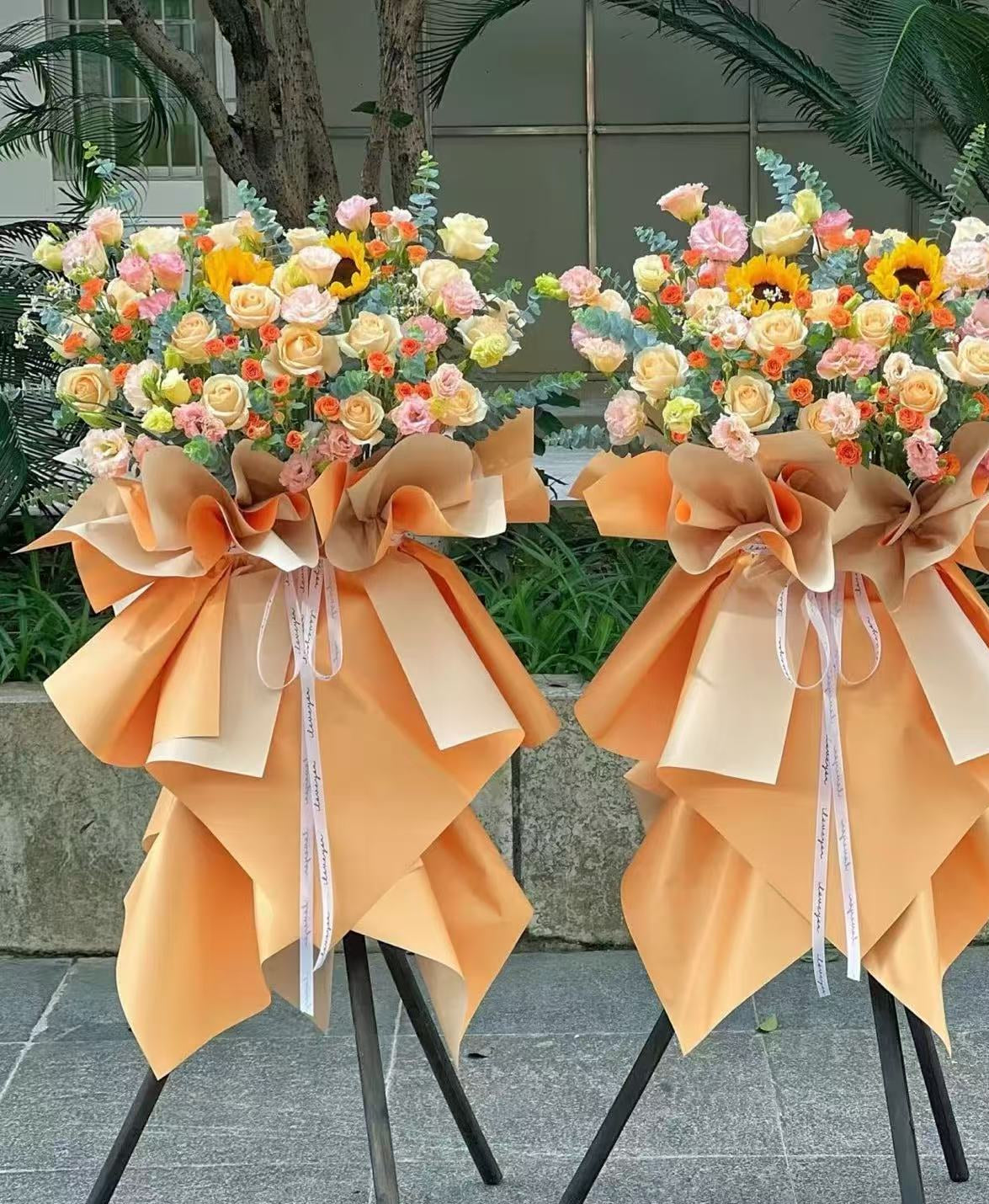 Two large floral greeting flower baskets on black metal stands, decorated with orange, yellow, pink, and white flowers and wrapped in peach and beige ribbon, set outdoors on a stone ledge.