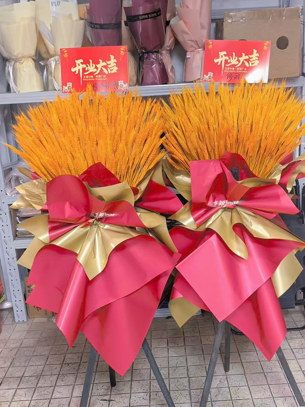 Two large decorative floral arrangements with bright orange-yellow flower stems, wrapped in red and metallic gold ribbon, displayed on stands in a shop setting. Red signs with Chinese characters are visible behind them.
